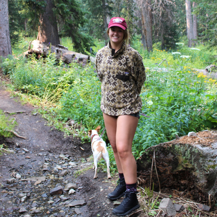Woman with a dog standing in a rocky forest trail with plants in the background.