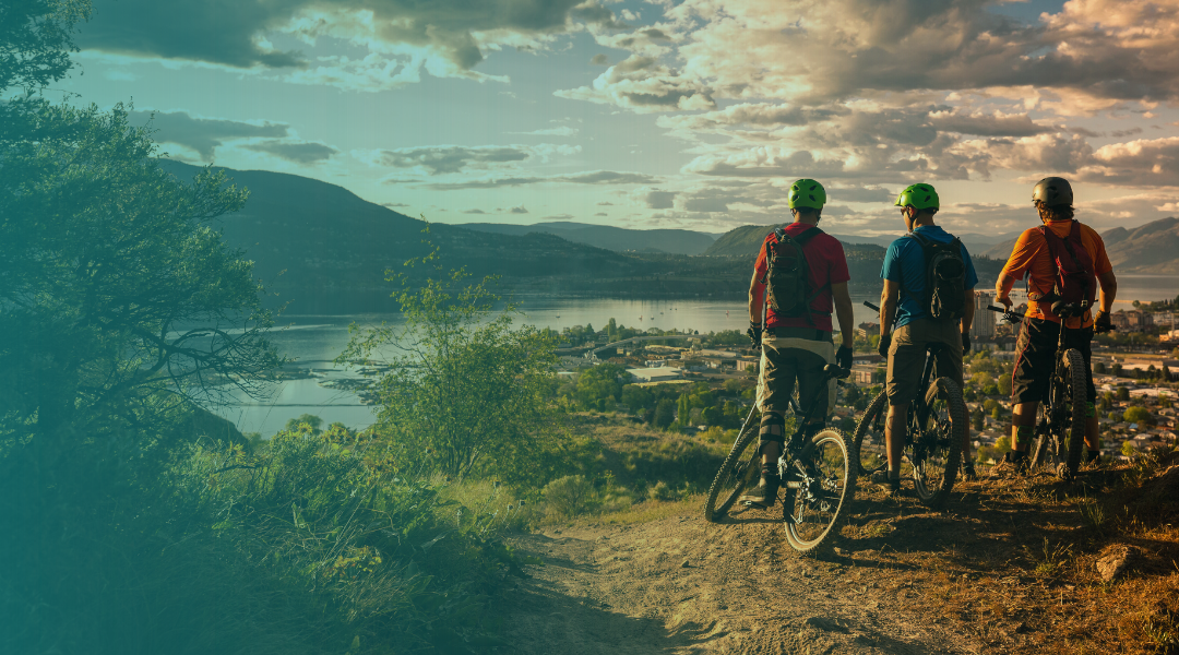 Three Grand Junction mountain bikers on a scenic trail with a lake and mountains in the background.