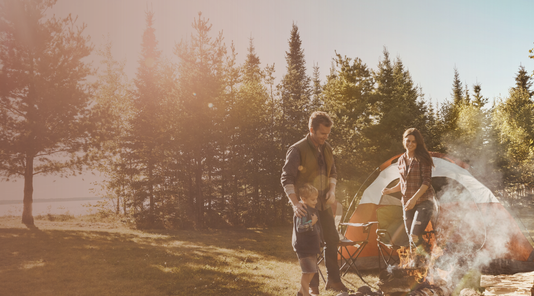 Two people and a child camping by a lake with a tent and fire pit.